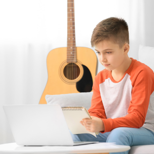 Child takes notes while looking at his laptop, a guitar hangs on the wall behind him.