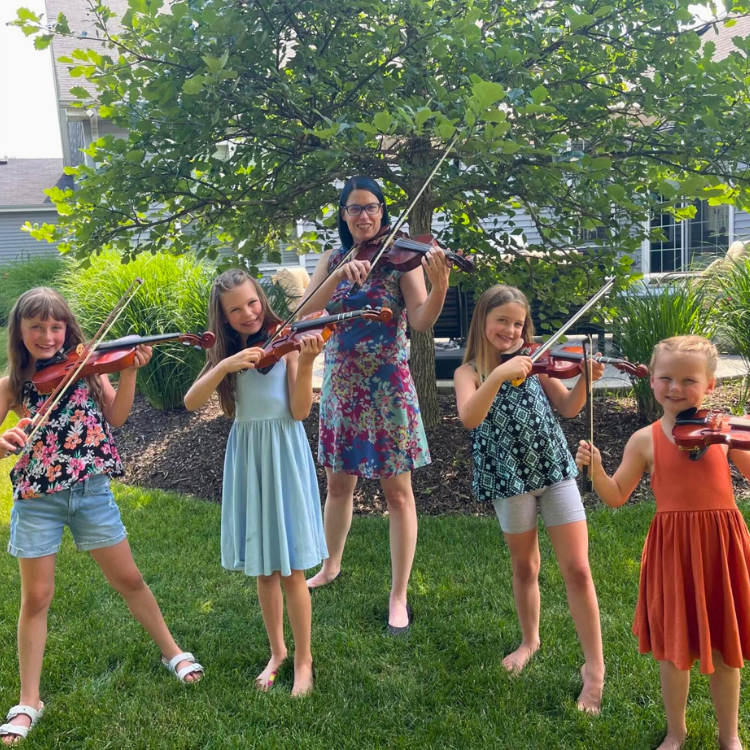 Stacey Sinclair posing outdoors for a photo with a group of girls who are learning violin. All are holding violins and bows in playing position and are smiling.