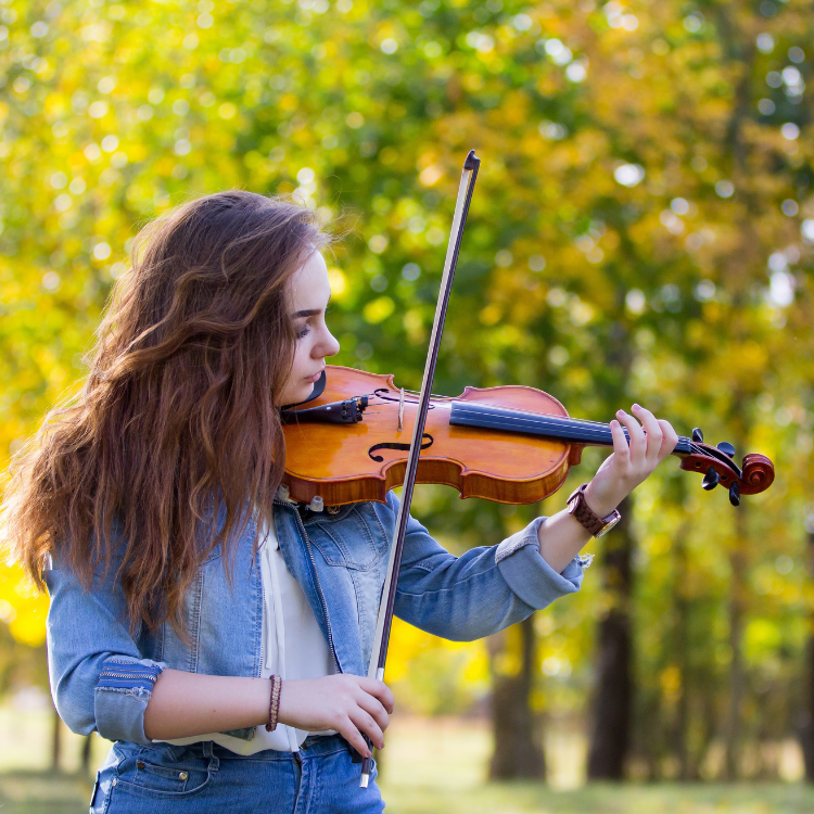 A young lady plays violin outdoors with green foliage in the background